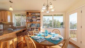 Een eetruimte met een houten tafel voor vier personen, omringd door stoelen, een boekenkast en een raam met uitzicht op bomen.