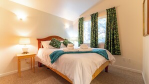 Een net opgemaakt bed met witte lakens, een houten hoofdbord en een blauwgroene plaid. Er staat een klein nachtkastje met een lamp en een stopcontact. De kamer heeft een beigekleurig tapijt en groene gordijnen bij het raam.