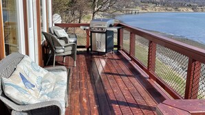 Una terraza de madera con sillas de mimbre y una parrilla, con vistas a un lago y montañas al fondo.