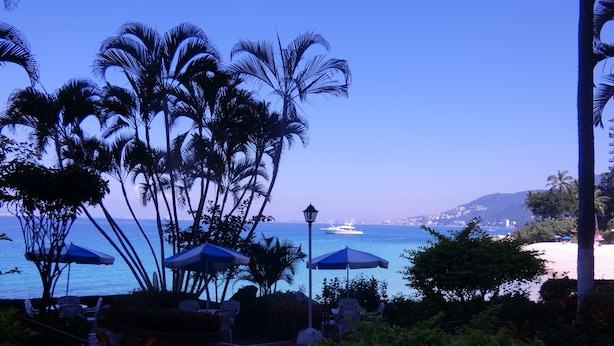 Un patio frente a la playa con sombrillas, palmeras y vista al océano y un yate.
