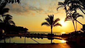 Un área de piscina con sillones de descanso y palmeras al atardecer.