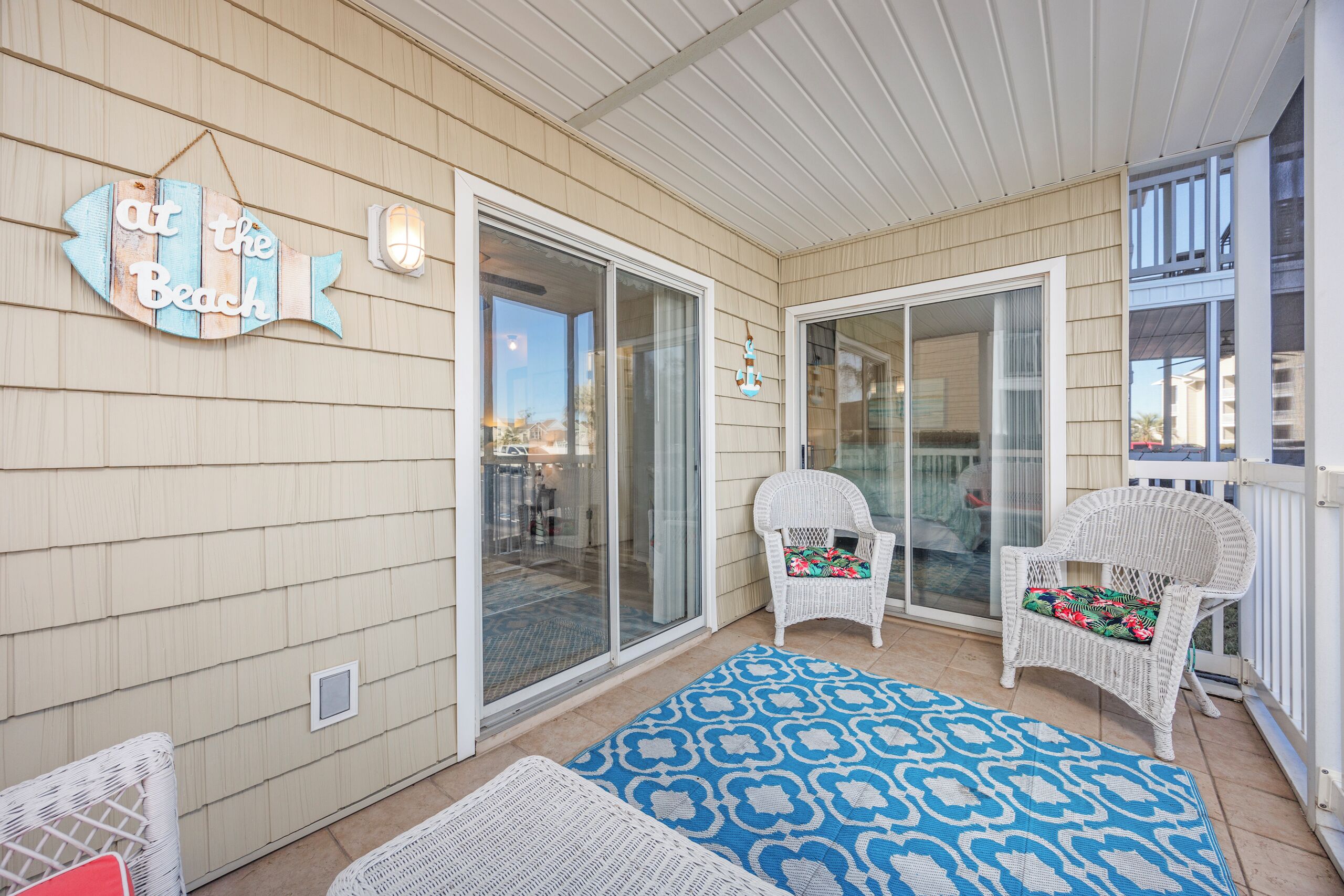 A covered outdoor area with a sliding glass door leading to a patio, two wicker chairs with colorful cushions, and a blue patterned rug.