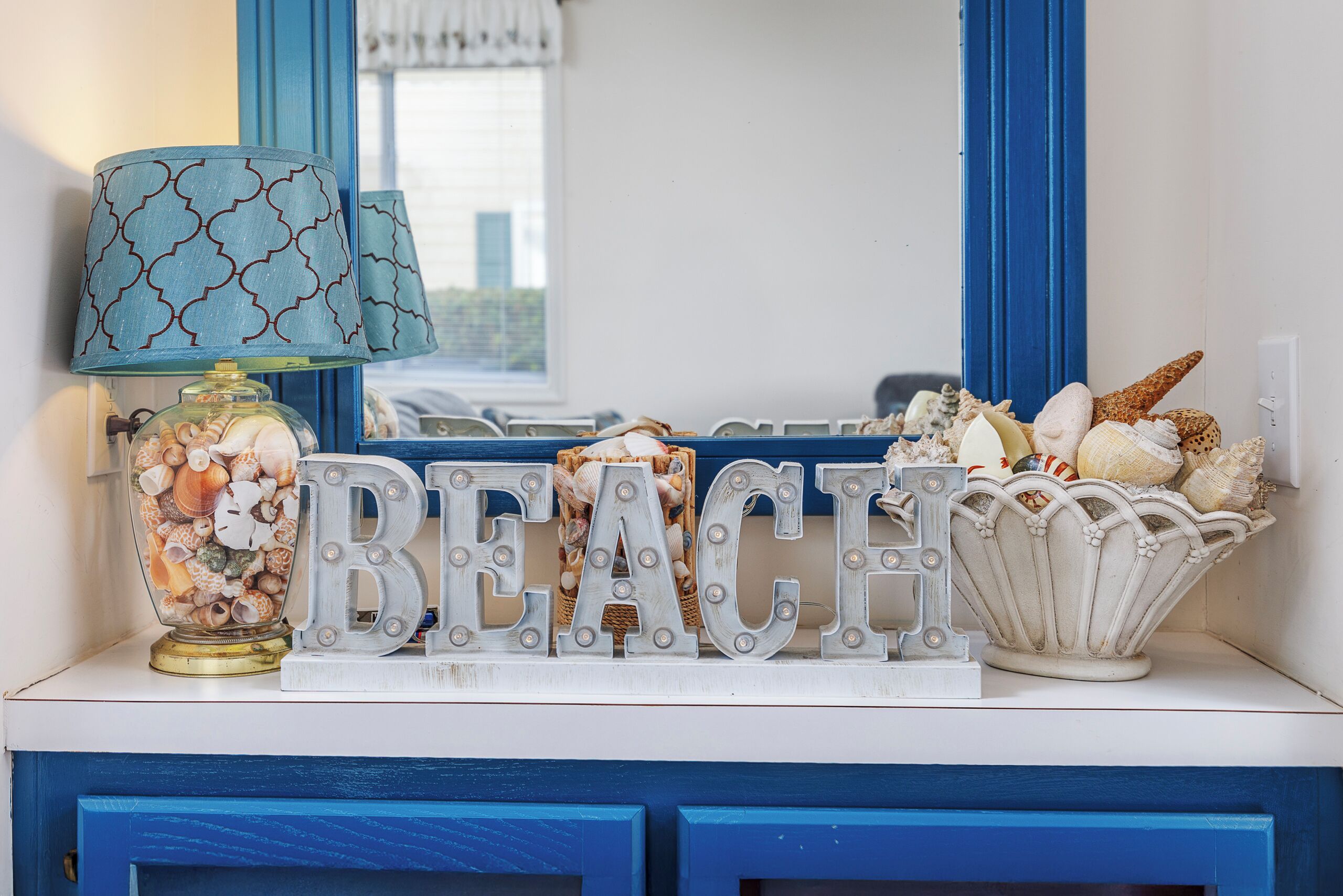 Eine dekorative Anordnung mit der leuchtenden Aufschrift „BEACH“, einer Lampe und einer Schale Meeresmuscheln.