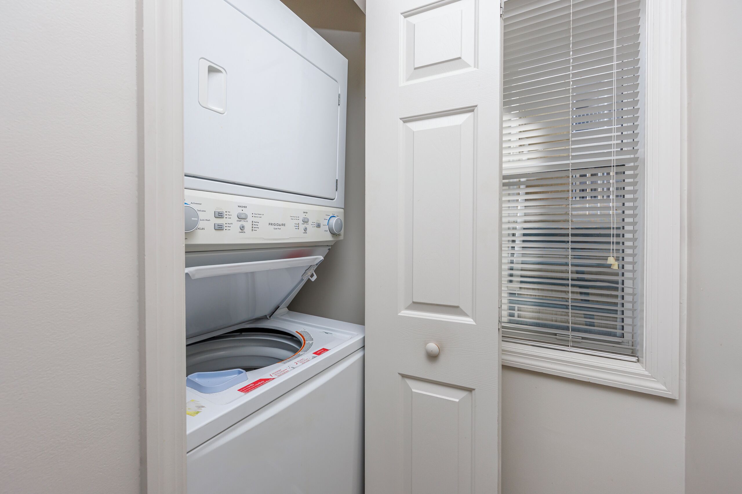 A compact laundry room with a front-loading washer and dryer, a door with a handle, and a window with blinds.