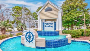 A pool area with a 'Waterway Landing' sign, surrounded by palm trees and a clear sky. - Affordable cozy 2 bedroom condo 1st floor screened in patio in gated community. (North Myrtle Beach)