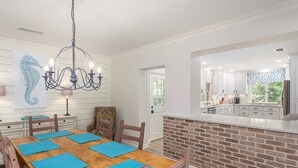 Uma sala de jantar com uma mesa de madeira posta para quatro pessoas, um candelabro e uma cozinha visível através de uma porta aberta.