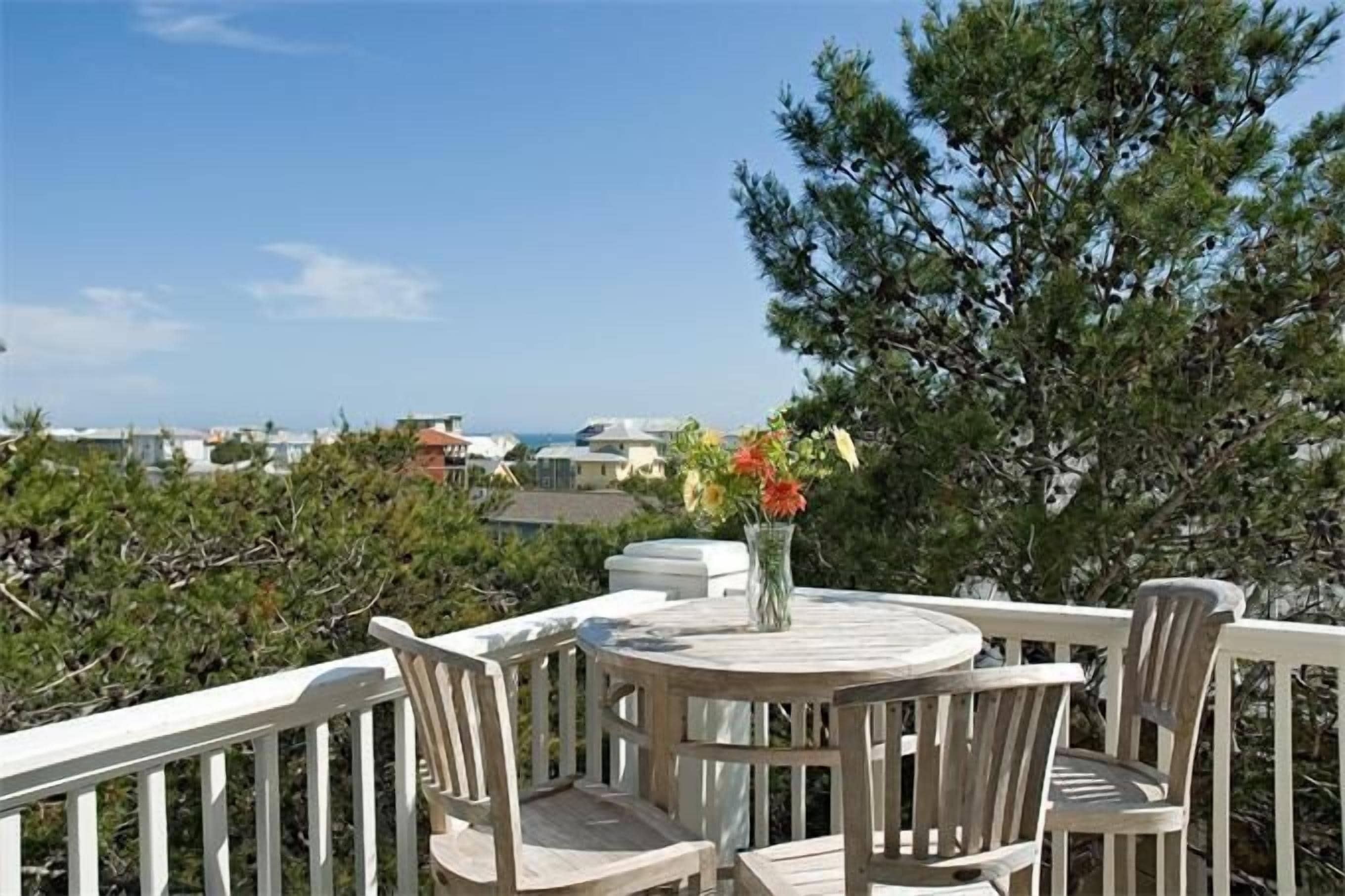 A round wooden table with four chairs, a vase with flowers, and a view of the ocean and buildings in the background.