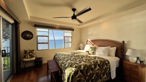 A bedroom with a large bed, a ceiling fan, a view of the sea through a window, a wooden nightstand, and a bench by the window. - Royal Mauian Oceanfront, Fabulous View!   (Kihei)