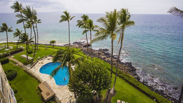 A pool area with a view of the ocean, surrounded by palm trees and a rocky shoreline. - Royal Mauian Oceanfront, Fabulous View!   (Kihei)