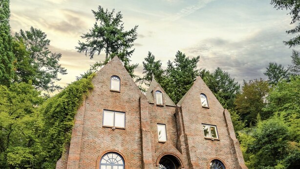 Ein Backsteinhaus mit mehreren Rundbogenfenstern und einer zentralen Tür, umgeben von üppiger Vegetation und einem Metallzaun.