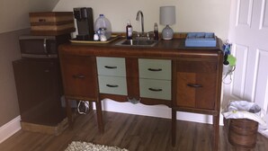 Cocina compacta con encimera de madera, un lavamanos y un microondas. Hay un cubo de basura pequeño y un armario de madera con cajones.