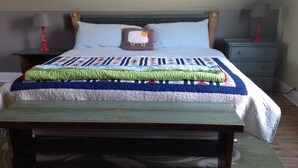 Una cama con colcha, cabecera de madera, mesita de noche con lámpara y estante fijado a la pared.