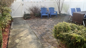 A paved patio with two blue Adirondack chairs, a wooden table, and a white fence with a lattice top.