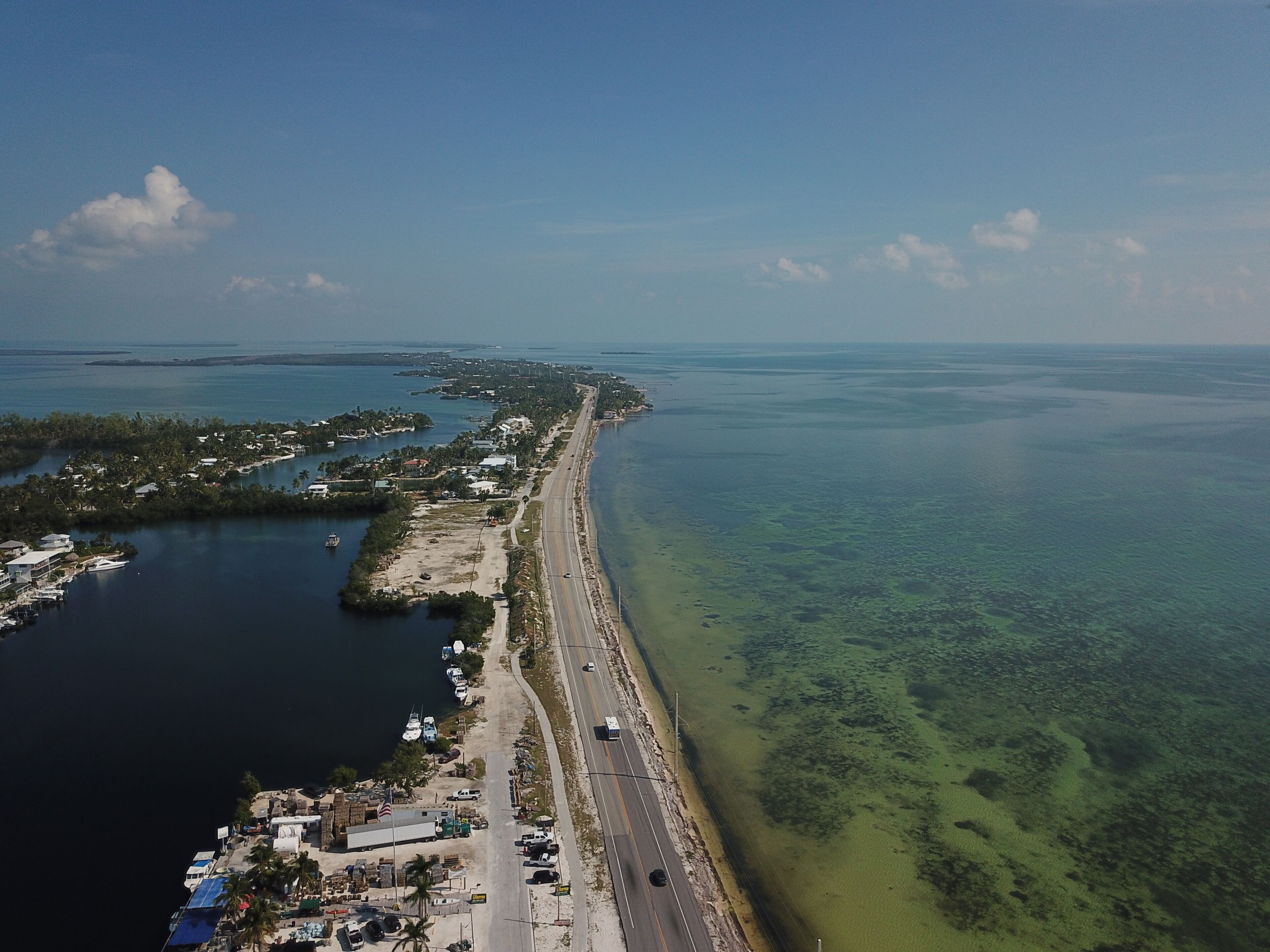 Une vue aérienne d’une zone côtière, avec une route qui longe la plage, des bateaux amarrés le long du rivage et une vue dégagée sur l’océan.