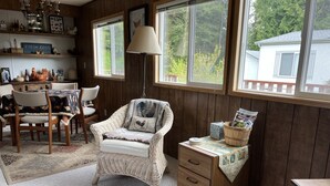 A cozy room with a wicker chair, a wooden side table with a lamp, and a view of a house through large windows.