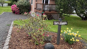 A small, single-story house with a deck, surrounded by a well-maintained garden with daffodils and a sign reading "Hen House".