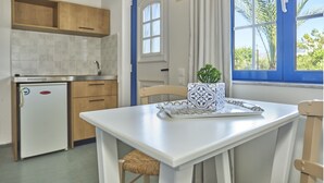 A compact kitchen with a small refrigerator, wooden cabinets, and a window with a view of palm trees. - Maistrali Sea View Apartments (Hersonissos)
