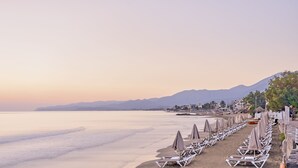 A beach with rows of sun loungers and umbrellas, a calm sea, and mountains in the background. - Maistrali Sea View Apartments (Hersonissos)