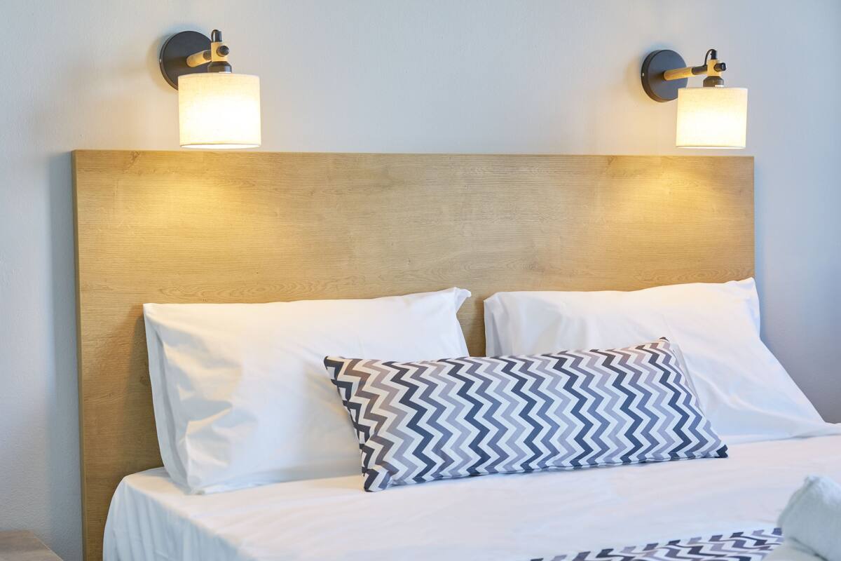 A hotel room with a wooden headboard, two white pillows, a patterned pillow, and a bedspread with a geometric design.