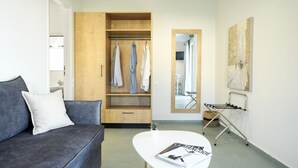 A modern living room with a grey sofa, a white coffee table with a magazine, a wooden wardrobe with hanging clothes, and a large mirror. - Maistrali Sea View Apartments (Hersonissos)
