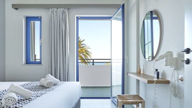 A hotel room with a bed, a small stool, a mirror, and a view of the balcony. - Maistrali Sea View Apartments (Hersonissos)