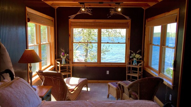 Una habitación acogedora con cama, una mesa con sillas, un ventanal con vistas al agua y un jarrón con flores.