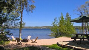 Un patio junto al lago con sillas Adirondack, una mesa de picnic y una sombrilla verde.