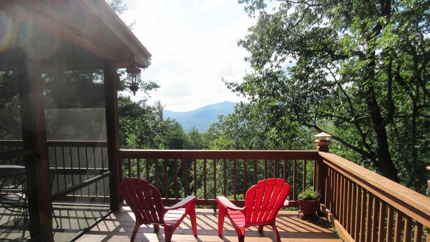 A wooden deck with two red chairs and a table, overlooking a forested area with mountains in the distance. - Entire cabin for rent, 30-day minimum stay.   (Clayton)