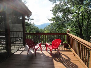 A wooden deck with two red chairs and a table, overlooking a forested area with mountains in the distance.