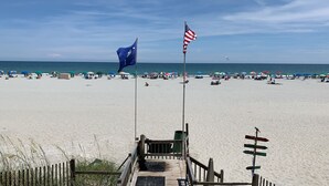 En strand med en träpromenad som leder ner till vattnet, med en flaggstång som bär en amerikansk flagga och en blå flagga.