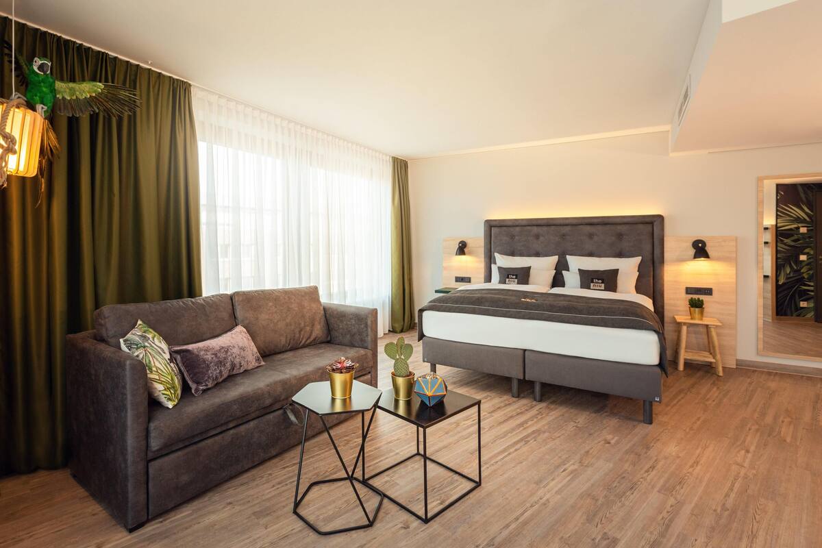 A modern hotel room with a bed, a sofa, a small table, and a large window with curtains.
