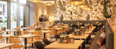 A restaurant with multiple tables and chairs, hanging pendant lights, and a wall with a leafy pattern.