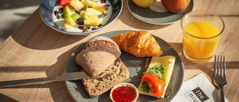 Un desayuno con croissants, queso, tomate, pan y un vaso de zumo de naranja sobre una mesa de madera.