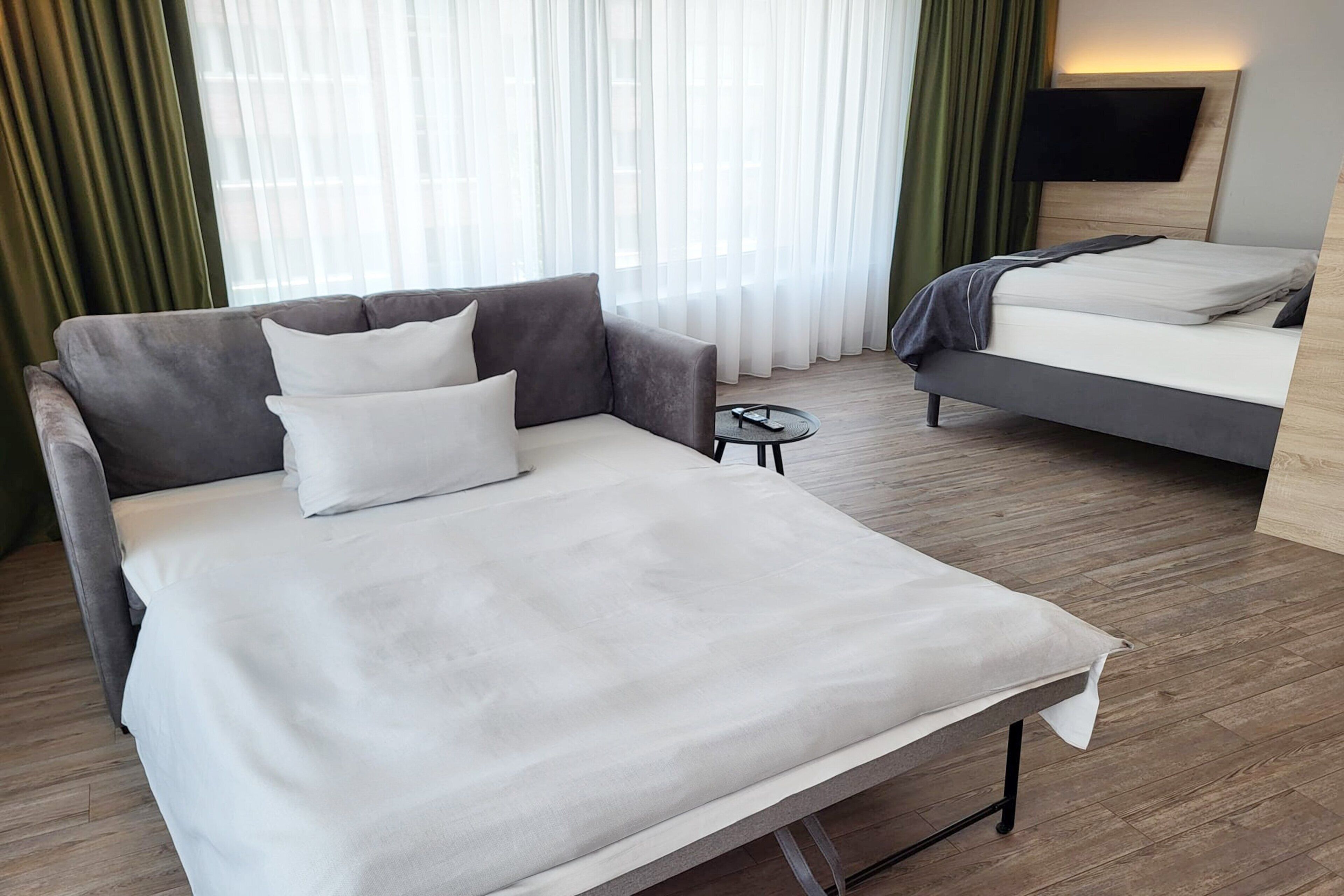 A modern hotel room with a grey sofa, a single bed, a small round table, and a TV mounted on the wall.