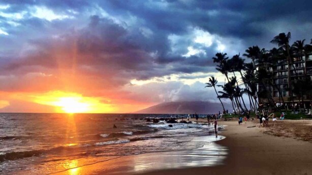 Ein Strand bei Sonnenuntergang, mit Palmen und einem Gebäude im Hintergrund.
