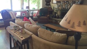 A cozy living room with a stone fireplace, comfortable seating, and a large window providing natural light. - Camp Pinecone--Waterfront Home on Upper Saranac Lake (Saranac Lake)