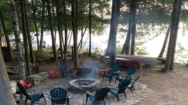 A stone fire pit surrounded by Adirondack chairs in a forested area with a view of the lake. - Camp Pinecone--Waterfront Home on Upper Saranac Lake (Saranac Lake)