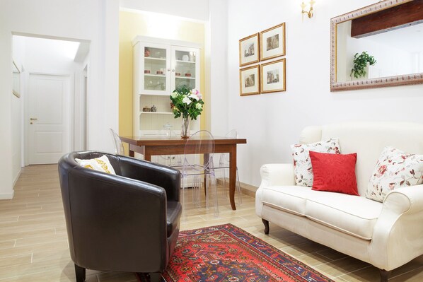 A living room with a leather armchair, a white sofa with red cushions, a dining table with clear chairs, a cabinet with glass doors, and a mirror on the wall.