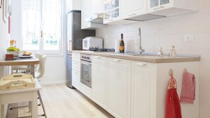 A modern kitchen with white cabinetry, a built-in oven, a microwave, and a gas stove. There is a window with lace curtains and a table with a bowl of fruit and a bottle of wine. - Beautiful, Bright, Spacious in Historic Bologna, great atmosphere, renovated (Bologna)