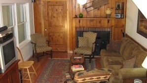 A cozy living room with wooden walls, a fireplace, a ceiling fan, and a television. - Charming Bear Paw Cabin with Spectacular View of Lake Hiwassee (Murphy)