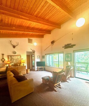 Un salón acogedor con techo de madera, chimenea, televisor y un ventanal que da a zonas verdes.