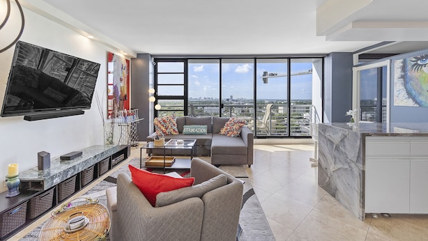 Ein modernes Wohnzimmer mit Sofa, Couchtisch und Fernseher, mit Blick auf die Stadt durch große Fenster.