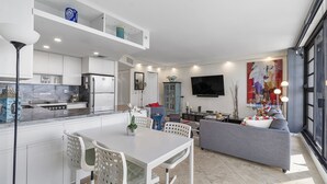 Eine moderne Küche mit weißen Schränken, eine Essecke mit weißem Tisch und Stühlen, ein graues Sofa und ein großes Fenster, das für natürliches Licht sorgt.