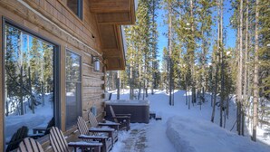 A wooden cabin with a patio area featuring wooden chairs and a hot tub, surrounded by snow and trees. -   Luxury Home, Ideal Winter Ski-in/Ski-out or Summer Basecamp (Big Sky)