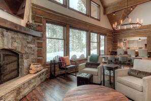 A log cabin interior with a stone fireplace, wooden floors, and large windows offering a view of the forest. -   Luxury Home, Ideal Winter Ski-in/Ski-out or Summer Basecamp (Big Sky)