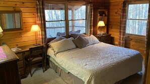A cozy log cabin bedroom with a bed, two bedside lamps, a desk with a chair, and a window with curtains.