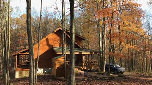 A cabin surrounded by trees with autumn foliage, a covered porch, and a car parked nearby.