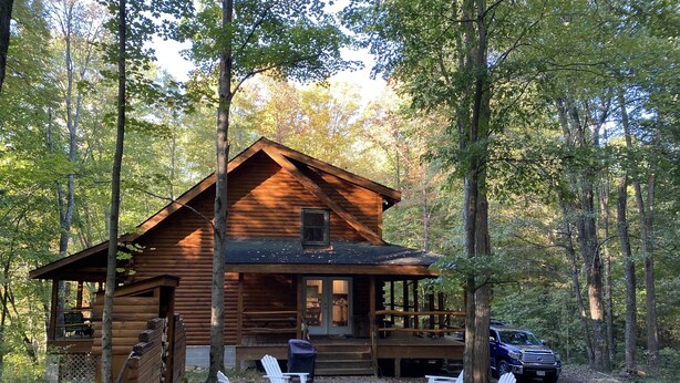 A log cabin with a porch, two Adirondack chairs, and a vehicle parked nearby.