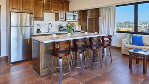 A modern kitchen with a central island, stainless steel refrigerator, wooden cabinets, and a large window offering a city view. - Beautiful condo located one block to Medano beach and The Cabo Marina (Cabo San Lucas)
