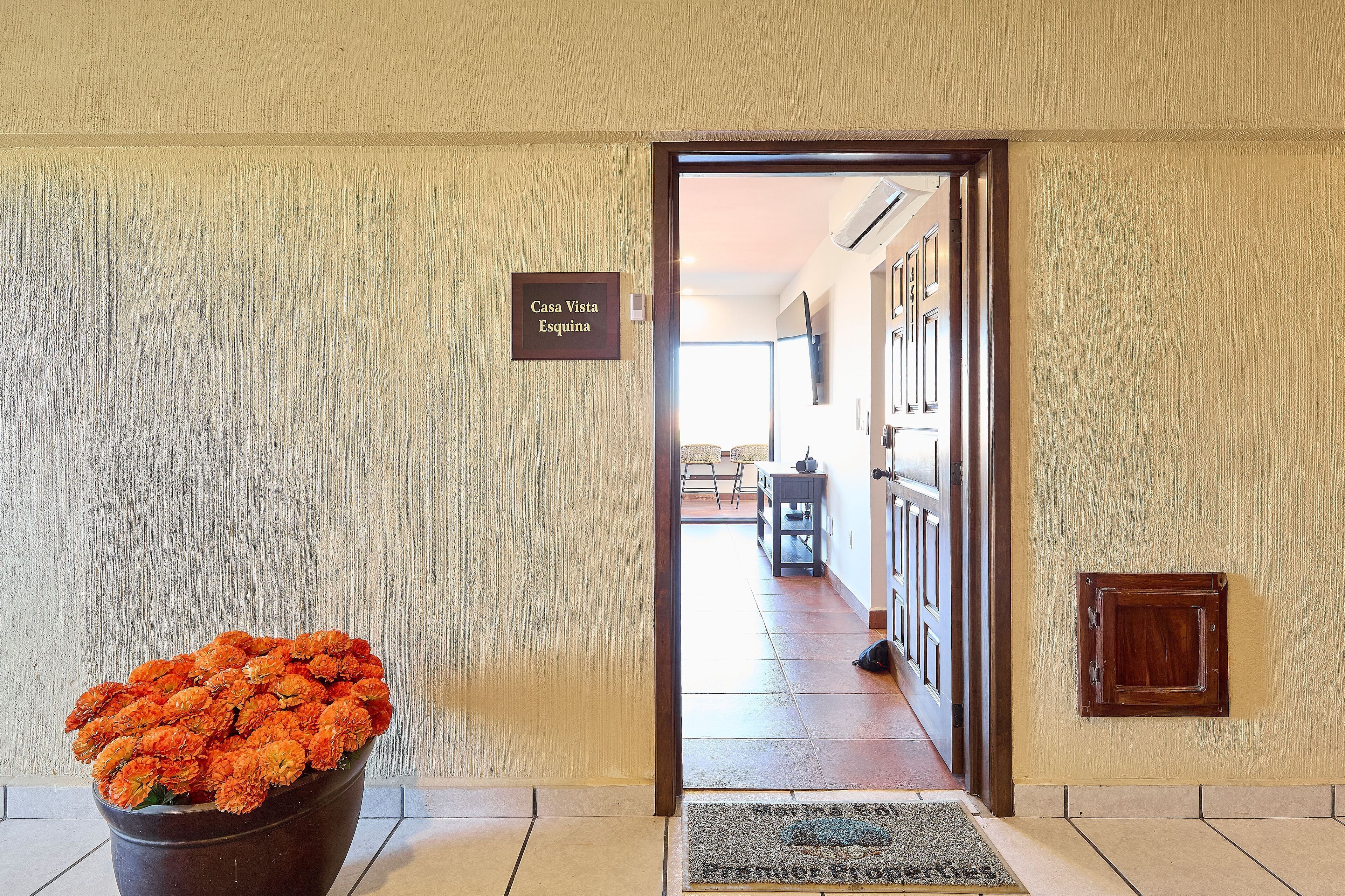 Entrada de hotel con un letrero que dice "Casa Vista Esquina", una planta en maceta con flores naranjas y un marco de puerta de madera.